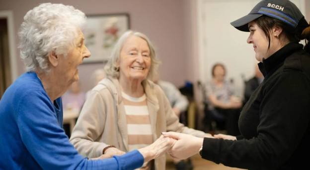 An elderly woman dancing with a caregiver in a senior living community.