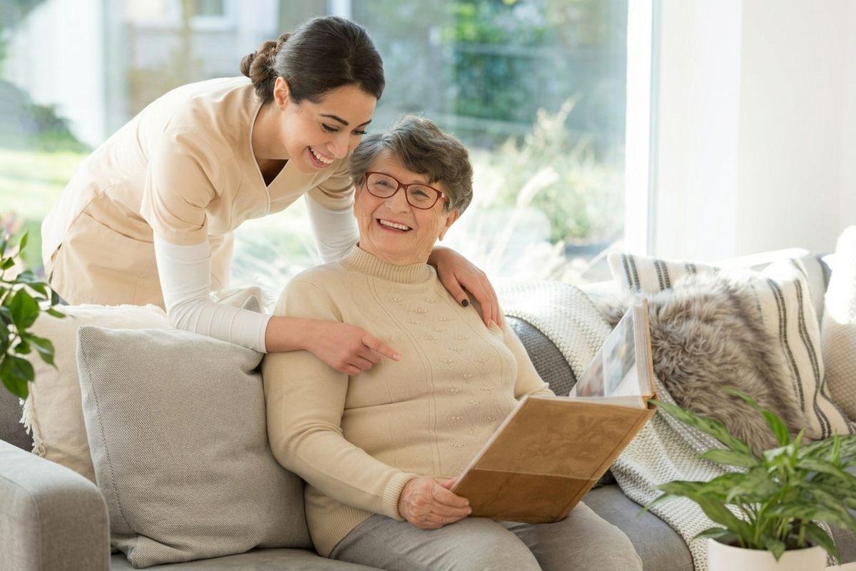 A caregiver leans over a smiling older adult on a sofa, both looking at a photo album by a bright window.